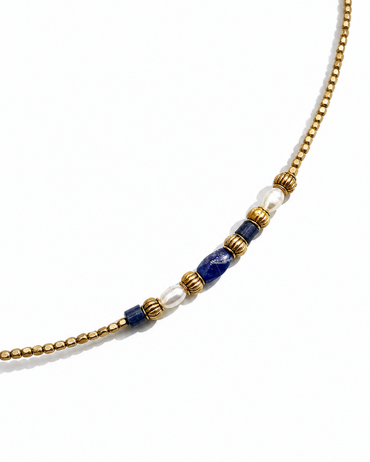 A gold beaded necklace featuring a central decorative cluster with white pearl-like beads, lapis lazuli-style blue beads, and textured gold spacer rings, shown on a clean white background.