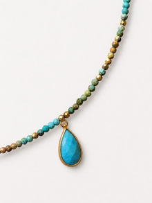 A beaded necklace featuring a large faceted turquoise teardrop pendant in a gold-tone bezel, attached to a strand of small round beads in turquoise, green, and bronze tones, photographed on a clean off-white background.